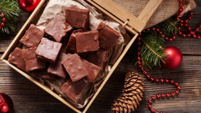 Box of chocolate fudge surrounded by holiday decor, including pinecones and ornaments, on wooden background.