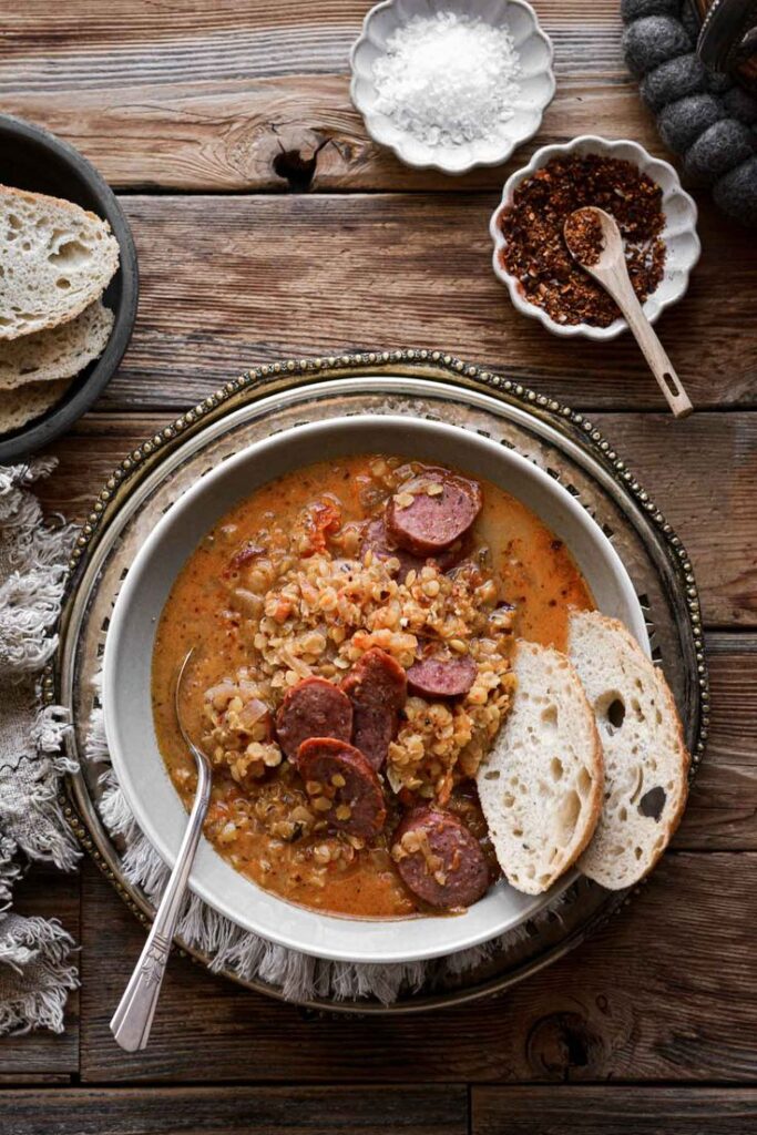 Hearty lentil and sausage stew with rustic bread on a wooden table, garnished with spices and salt in bowls.