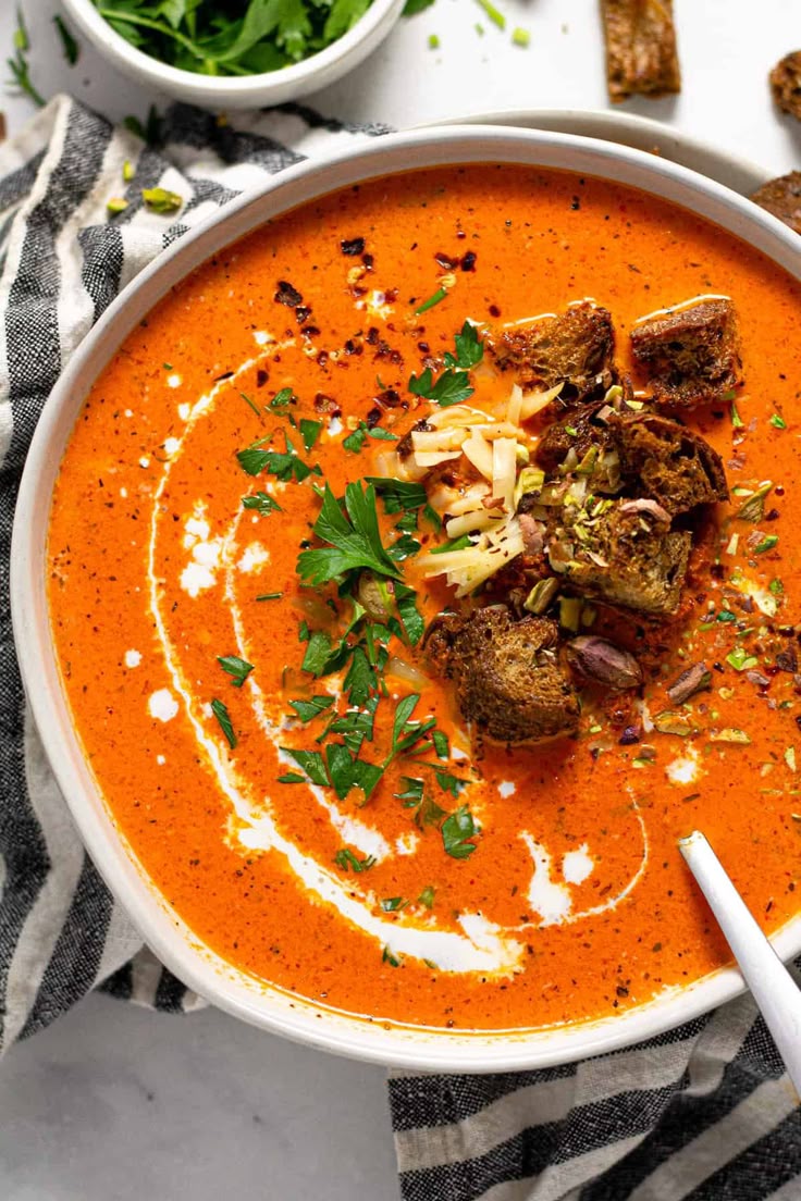 Creamy roasted tomato soup garnished with croutons, herbs, and cheese in a white bowl.
