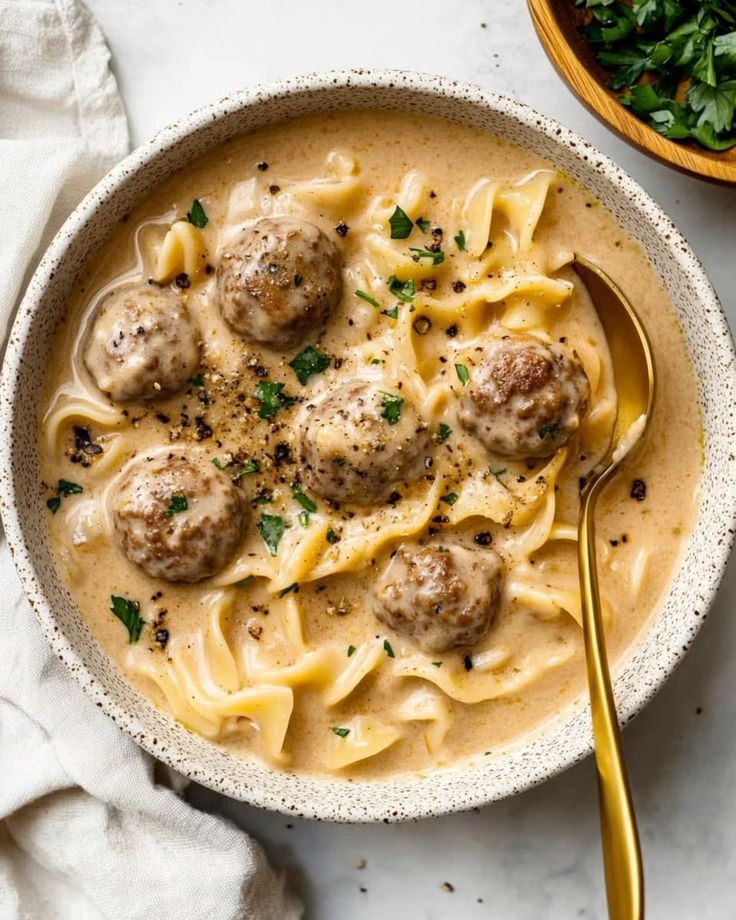 Creamy meatball pasta with parsley garnish in a ceramic bowl, gold spoon, and white napkin on the side.