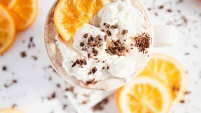 Creamy hot chocolate topped with whipped cream, chocolate shavings, and orange slices on a white background.