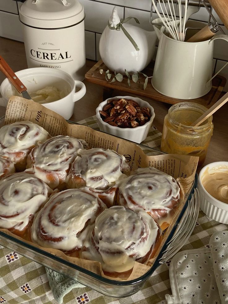 Freshly baked cinnamon rolls with creamy icing in a kitchen setting with nuts and yellow spread nearby.