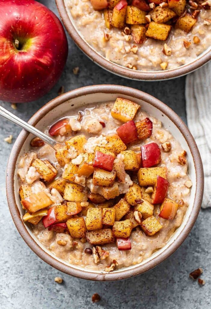 Apple cinnamon oatmeal is perfectly creamy and - Easy Christmas Breakfast Recipes for Joyful Mornings Bowl of oatmeal topped with caramelized apple chunks and pecans, next to a fresh whole apple on a gray background.
