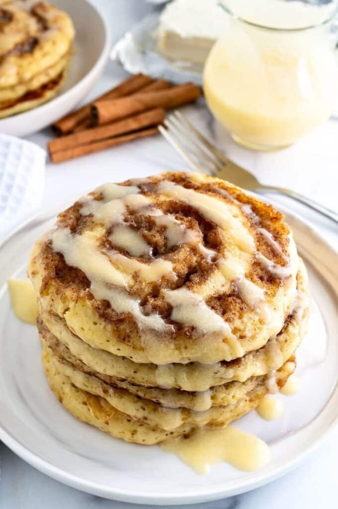 Cinnamon Roll Pancake Recipe - Easy Christmas Breakfast Recipes for Joyful Mornings Stack of cinnamon roll pancakes drizzled with icing, set beside cinnamon sticks and cream, on a white plate.