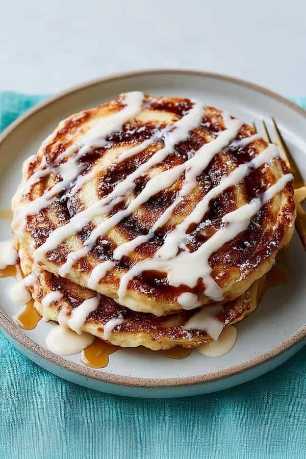 Cinnamon Roll Pancakes Recipe - SweetNourish Desserts for Every Lifestyle - Easy Christmas Breakfast Recipes for Joyful Mornings Cinnamon roll pancakes drizzled with icing on a plate, served on a blue tablecloth for breakfast.