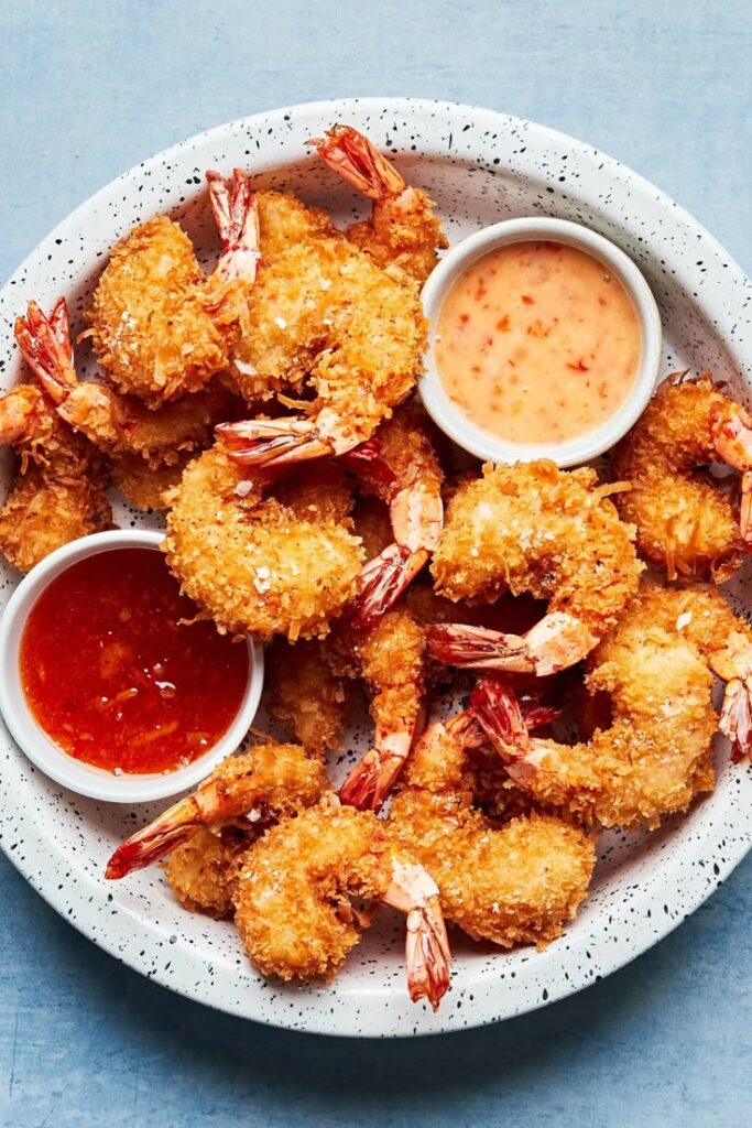 Coconut Shrimp - Crispy Coconut Shrimp & Sweet Chili Sauce Recipe Crispy fried shrimp with breadcrumbs served with two dipping sauces on a speckled white plate.