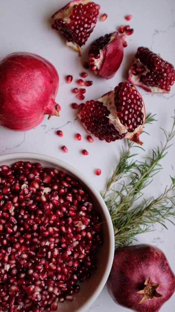 Delicious homegrown Pomegranates - Festive & Easy: Master the Perfect Pomegranate Paloma Fresh pomegranate seeds in a bowl with whole and halved pomegranates on a white background with rosemary sprigs.