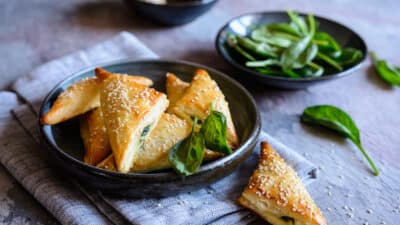 Golden spinach and cheese triangles topped with sesame seeds on a plate, with fresh spinach in the background.
