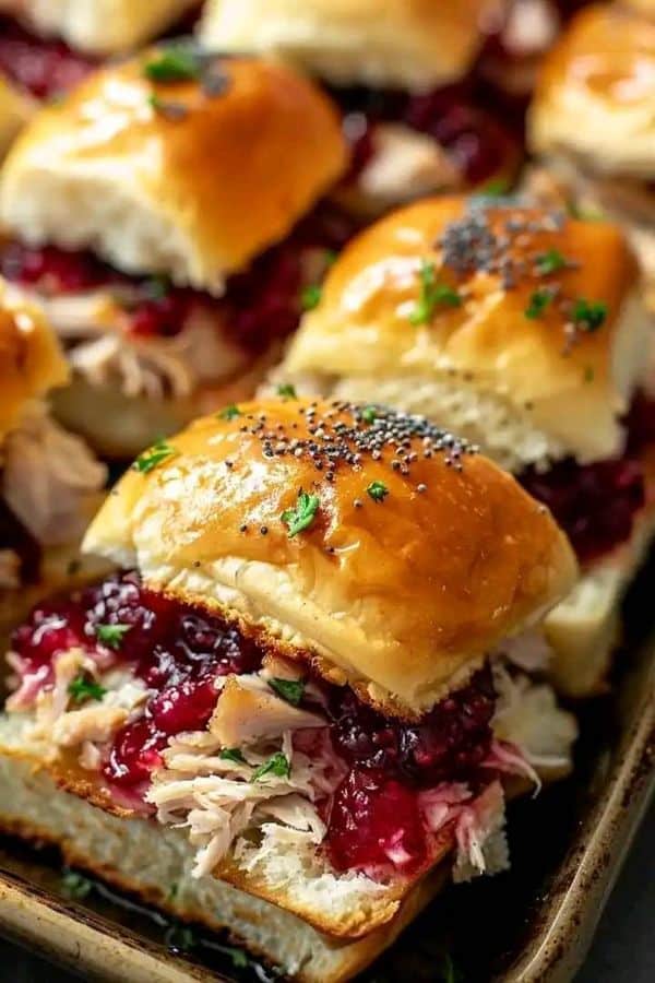 Sliders with turkey, cranberry sauce, and poppy seed-topped buns, garnished with parsley on a baking sheet.