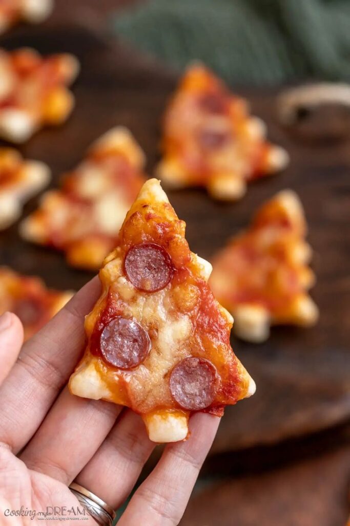 Mini Christmas tree-shaped pizza appetizers with pepperoni toppings held in hand.