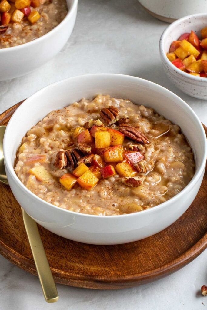 Slow Cooker Apple Cinnamon Oatmeal - Easy Christmas Breakfast Recipes for Joyful Mornings Bowl of oatmeal topped with diced peaches and pecans, served on a wooden platter with a golden spoon.