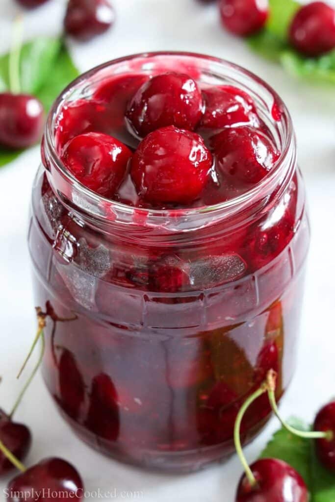 Homemade cherry preserves in a glass jar surrounded by fresh cherries on a white surface.