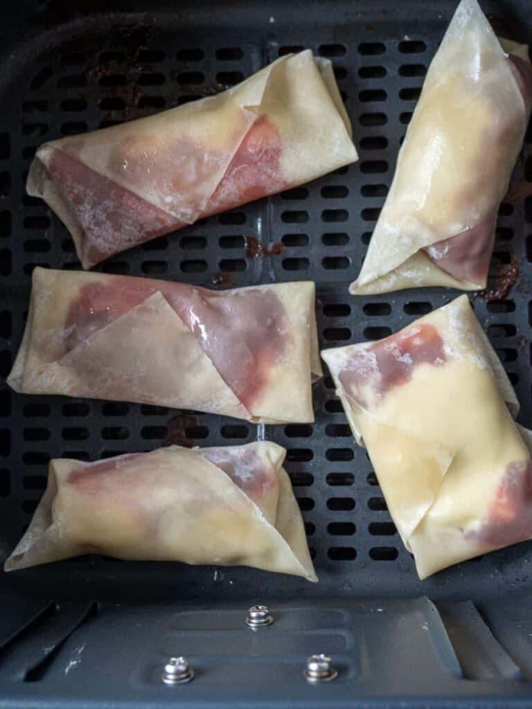 Five egg rolls in an air fryer basket, ready for cooking.