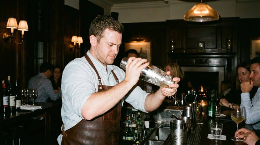 image - Blueberry Snow Martini: Cozy Winter Cocktail Recipe Bartender mixing cocktail at a cozy bar with patrons in the background.