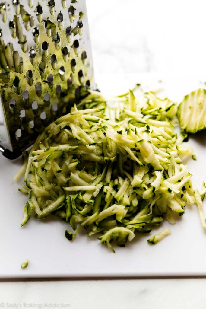 shredded-zucchini-with-box-grater - Versatile Zucchini Bread Recipe: Nutty, Chocolate, or Spiced Grated zucchini on white board with metal grater, ideal for baking or cooking recipes.