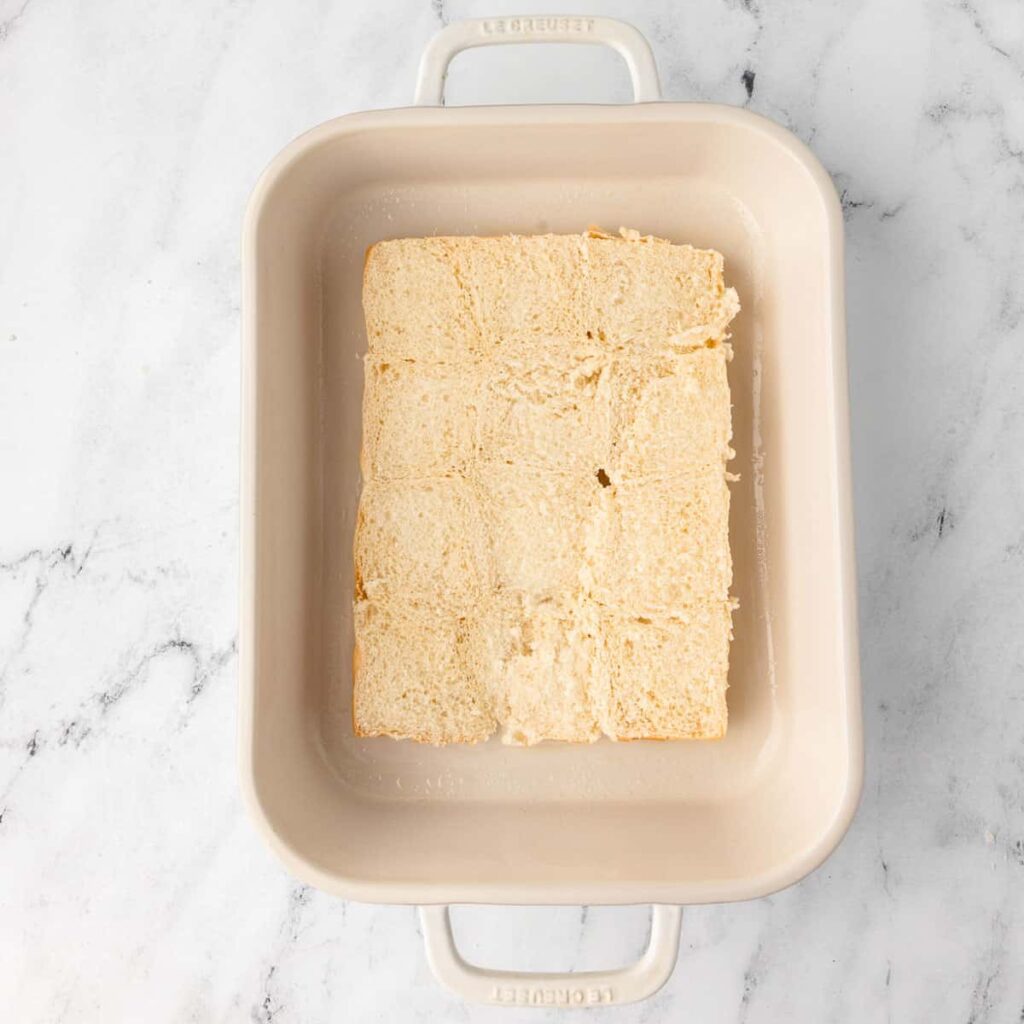 Cubed bread in a cream baking dish on a marble surface, ready for a casserole or bread pudding.