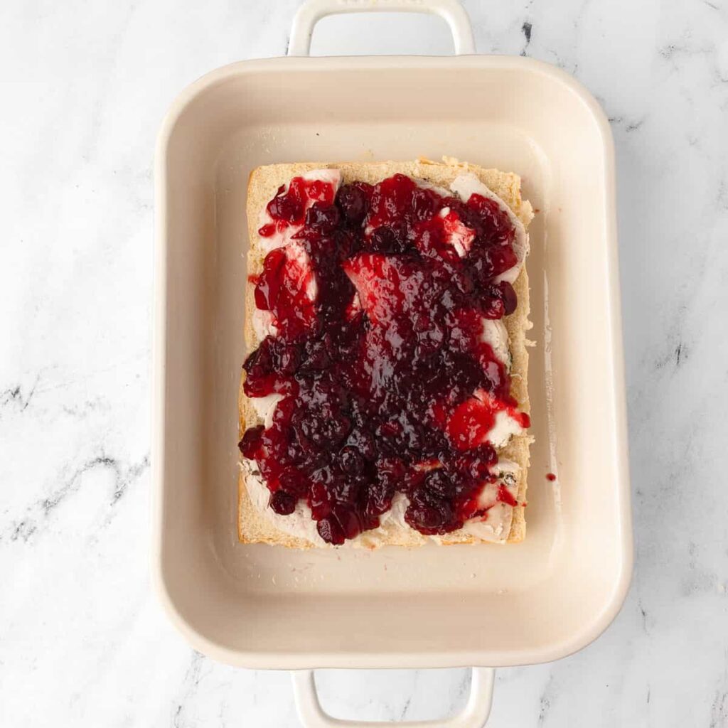 Bread slice topped with creamy spread and vibrant berry jam in a ceramic baking dish on a marble surface.