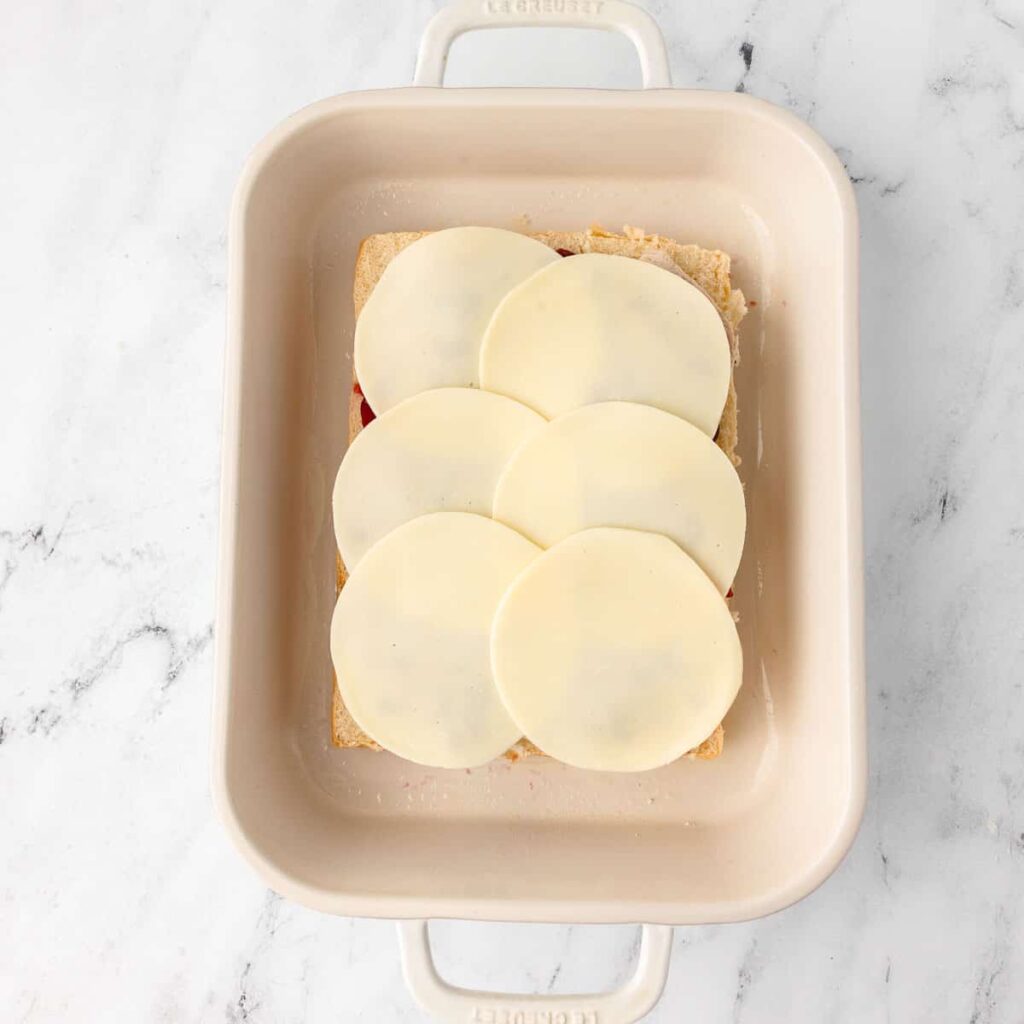 Baking dish with layered provolone cheese over lasagna on a white marble countertop.