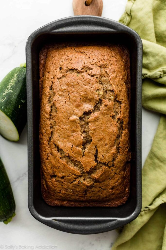 zucchini-bread-in-loaf-pan - Versatile Zucchini Bread Recipe: Nutty, Chocolate, or Spiced Freshly baked zucchini bread in a loaf pan, with green cloth and zucchinis on a marble countertop.