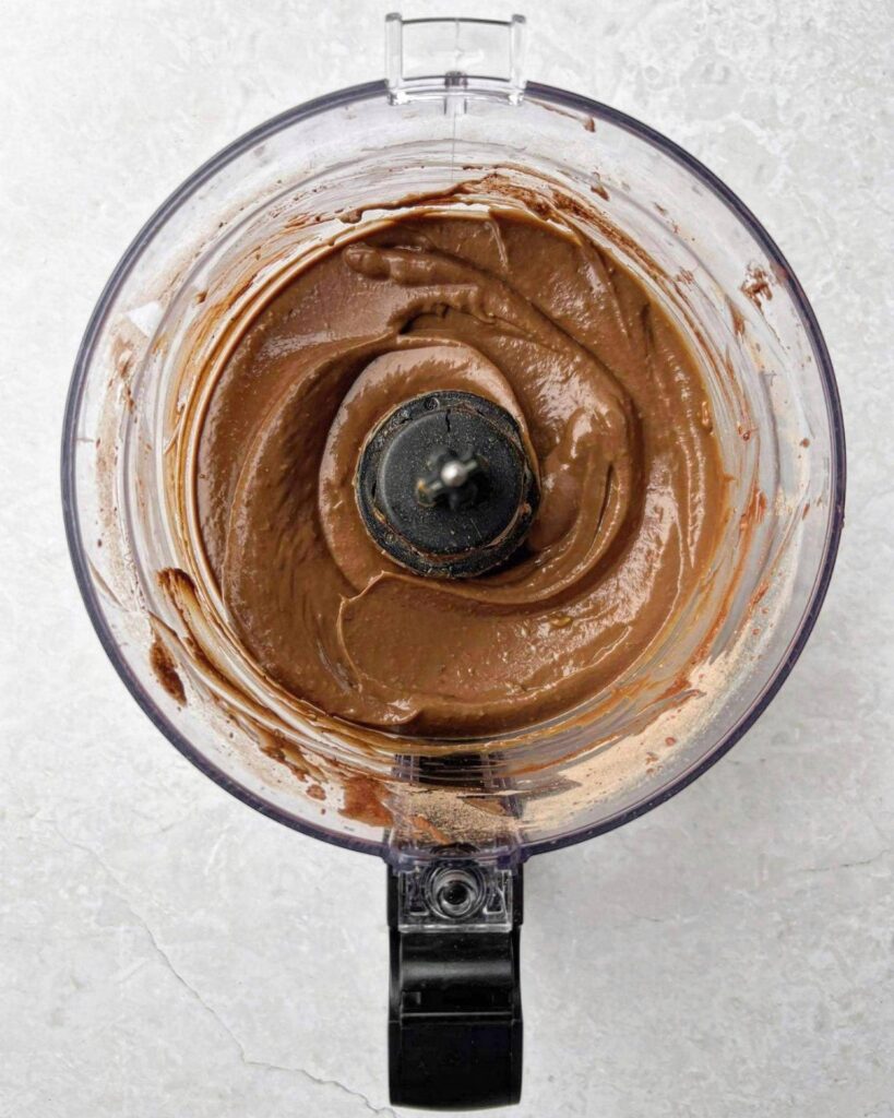 Chocolate spread being blended in a food processor for a smooth, creamy texture on a light grey countertop.