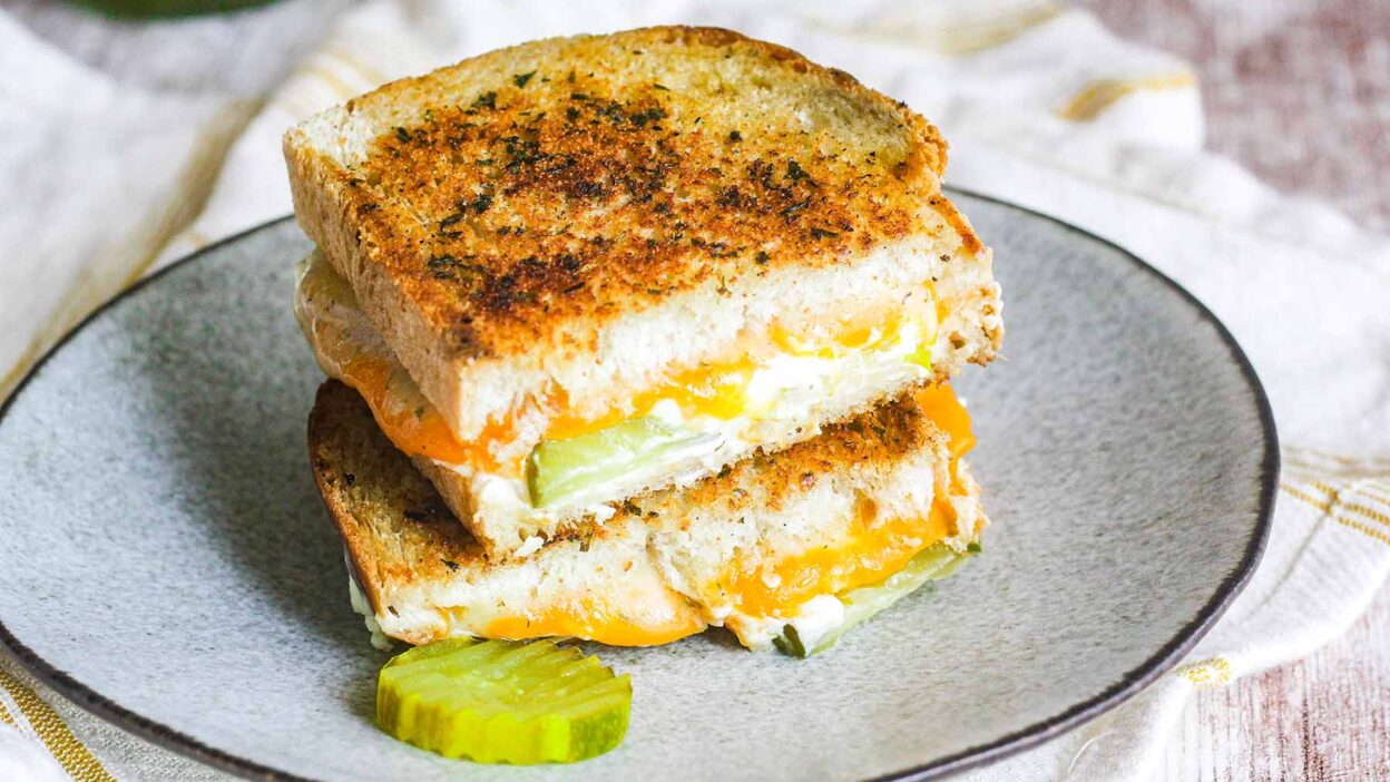 Grilled cheese sandwich with cheddar and pickles on a plate, showcasing a crispy golden crust.