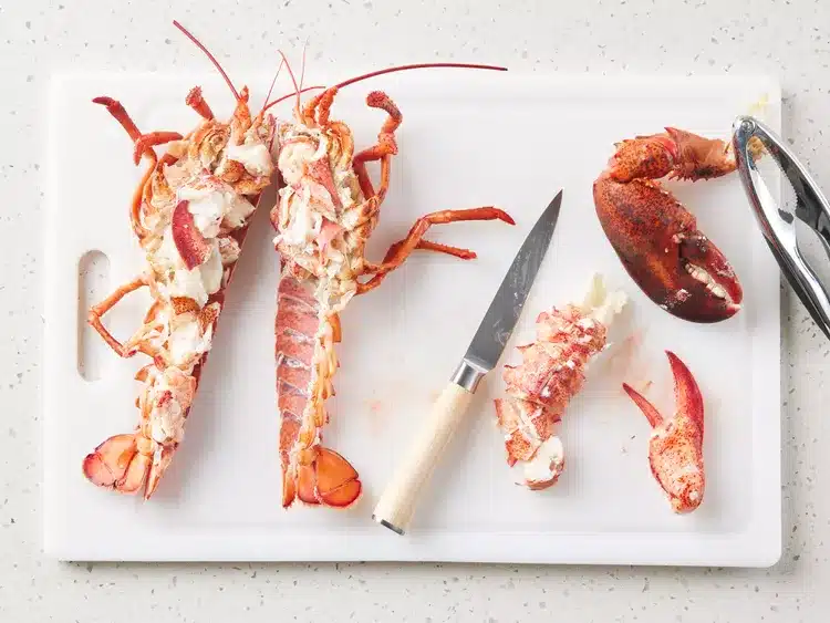 Fresh lobster cut in half on a white cutting board with knife and claw cracker nearby.