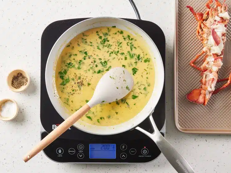 Creamy sauce cooking in a pan with herbs, alongside a prepared lobster on a tray, ready for serving.