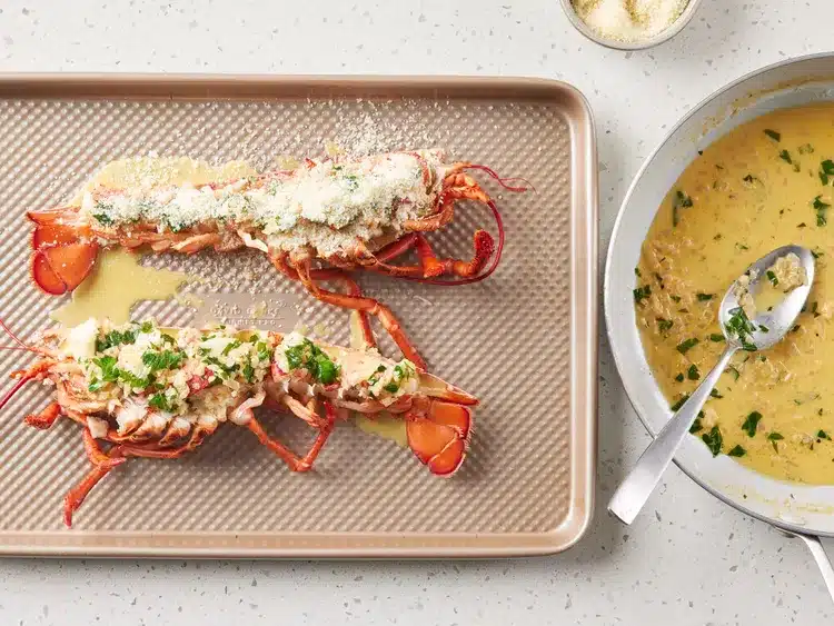 Two cooked lobsters with herbs and cheese on a baking tray, next to a pan with creamy sauce.