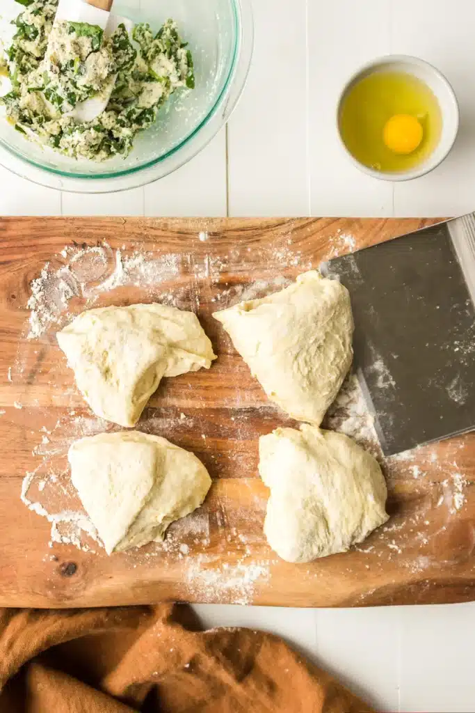 Four dough pieces, egg, spinach filling on wooden board, white background. Perfect for pastry preparation.