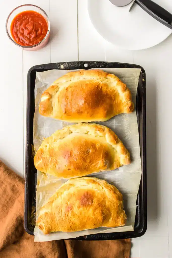 Three golden-brown empanadas on a baking sheet with marinara sauce and a pizza cutter on the side.