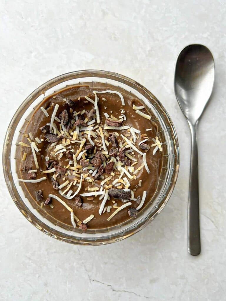Chocolate pudding topped with shredded coconut and cocoa nibs in a glass bowl with a spoon beside it on a marble table.