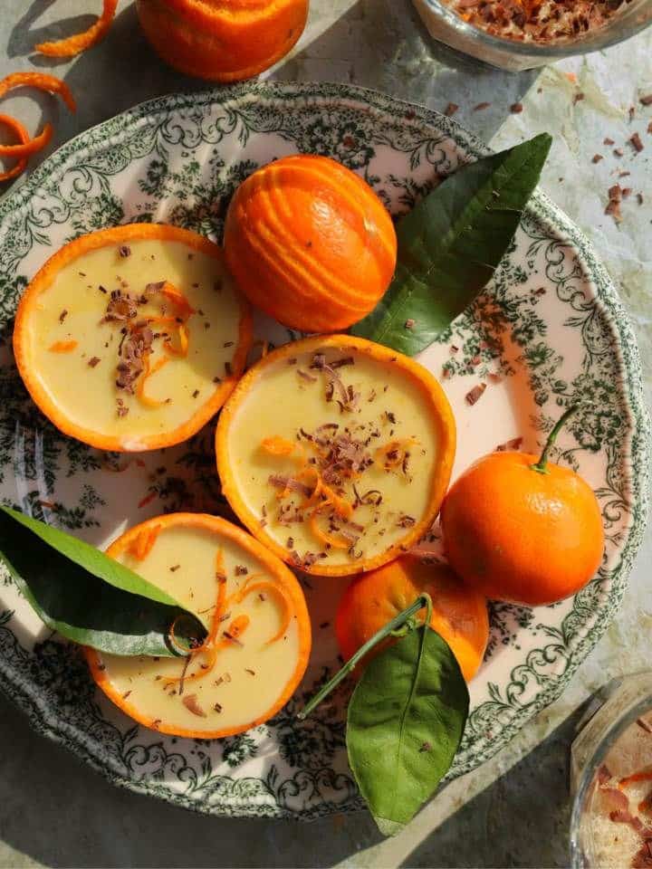 Creamy orange dessert cups garnished with chocolate shavings on a decorative plate with whole oranges and leaves.