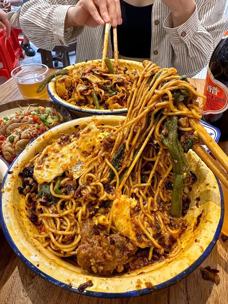 Person enjoying spicy Asian noodles with chopsticks, served with a side dish and drink.