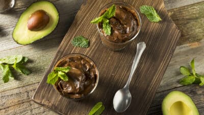 Creamy avocado chocolate mousse with mint garnish on a rustic board, surrounded by fresh avocados and mint leaves.