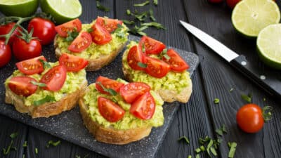 Avocado bruschetta topped with sliced tomatoes on a black plate, next to limes and tomatoes on a wooden table.