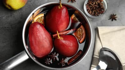 Pears poached in red wine with cinnamon and orange slices in a pot on a gray surface.