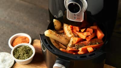 Air fryer with seasoned sweet potato and potato fries, herbs and spices on a wooden board.