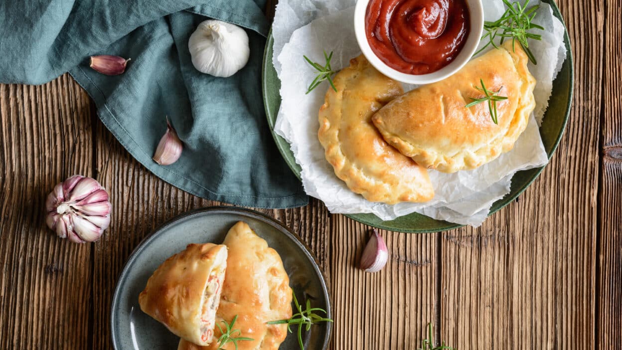 Delicious homemade empanadas with rosemary and dipping sauce on a wooden table, accompanied by garlic cloves.