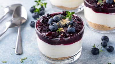 Blueberry cheesecake parfait in a glass with fresh blueberries and mint garnish on a light surface, featuring spoons nearby.