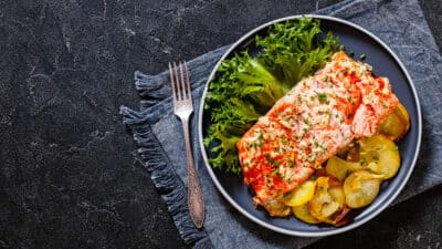 Grilled salmon with herbs, lettuce, and sliced potatoes on a blue plate and rustic napkin, top view, dark background.