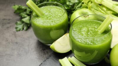 Two glasses of refreshing green smoothie with celery sticks, surrounded by fresh apples and leafy greens on a dark surface.