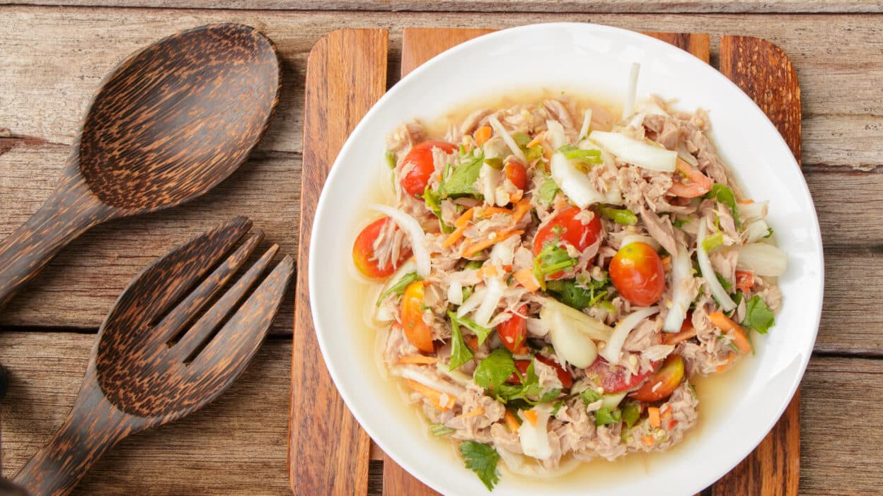 Fresh tuna salad with tomatoes, onions, and herbs in a white bowl on a wooden table with rustic cutlery.