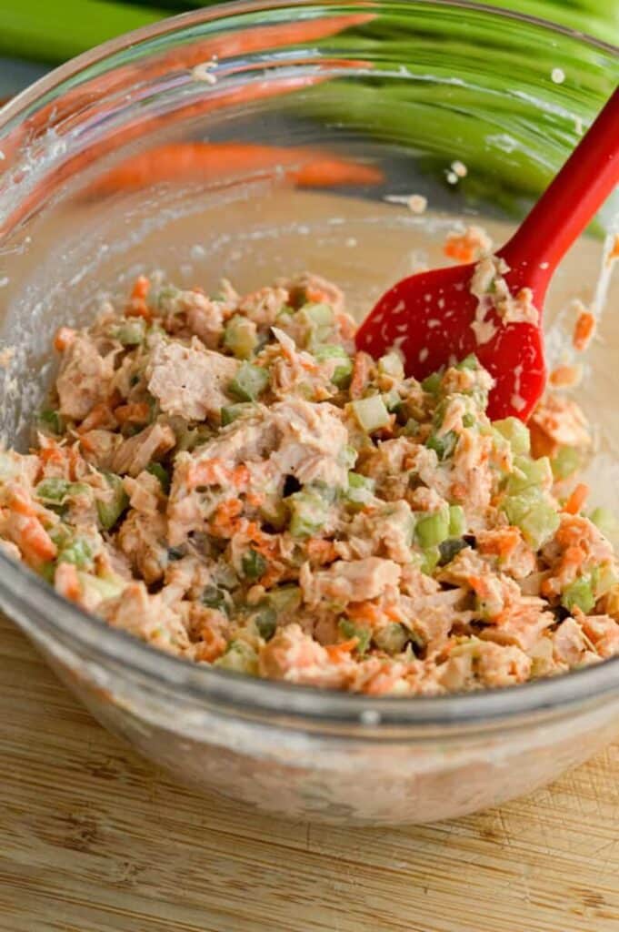 Homemade tuna salad mix with celery and carrots in a glass bowl, stirred with a red spatula on a wooden surface.