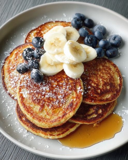 Fluffy pancakes topped with banana slices, blueberries, and syrup, sprinkled with powdered sugar on a plate.