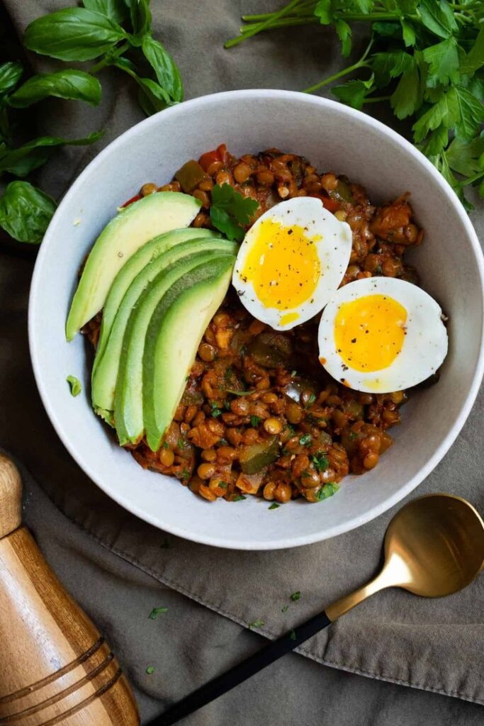 Bowl of spicy lentils with sliced avocado and soft-boiled eggs, garnished with herbs on a cloth napkin with spoon. High-Protein Breakfast 2026