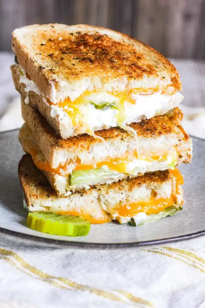 Stacked grilled cheese sandwiches with pickles on a plate, showcasing melted cheese and toasted bread.