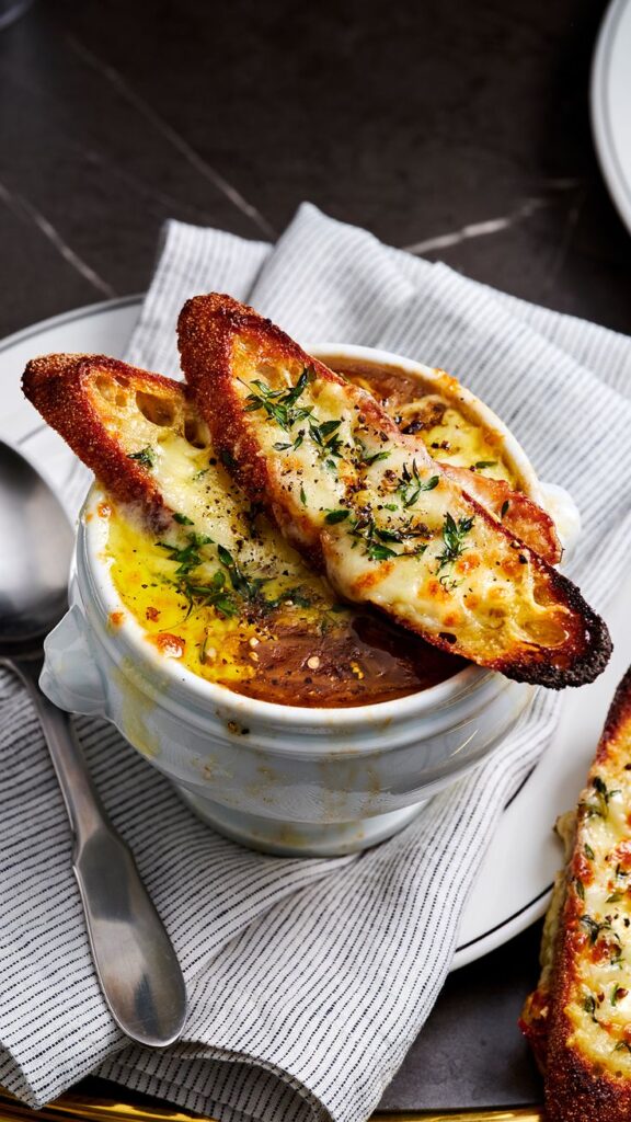 French onion soup topped with melted cheese and herbs on toast, served in a ceramic bowl with a spoon.