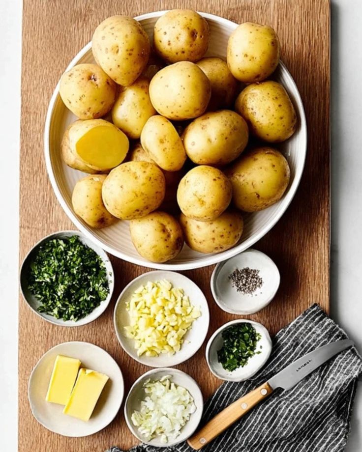 Bowl of potatoes with ingredients: butter, garlic, herbs, onions, and pepper, ready for a delicious recipe on a wooden board.