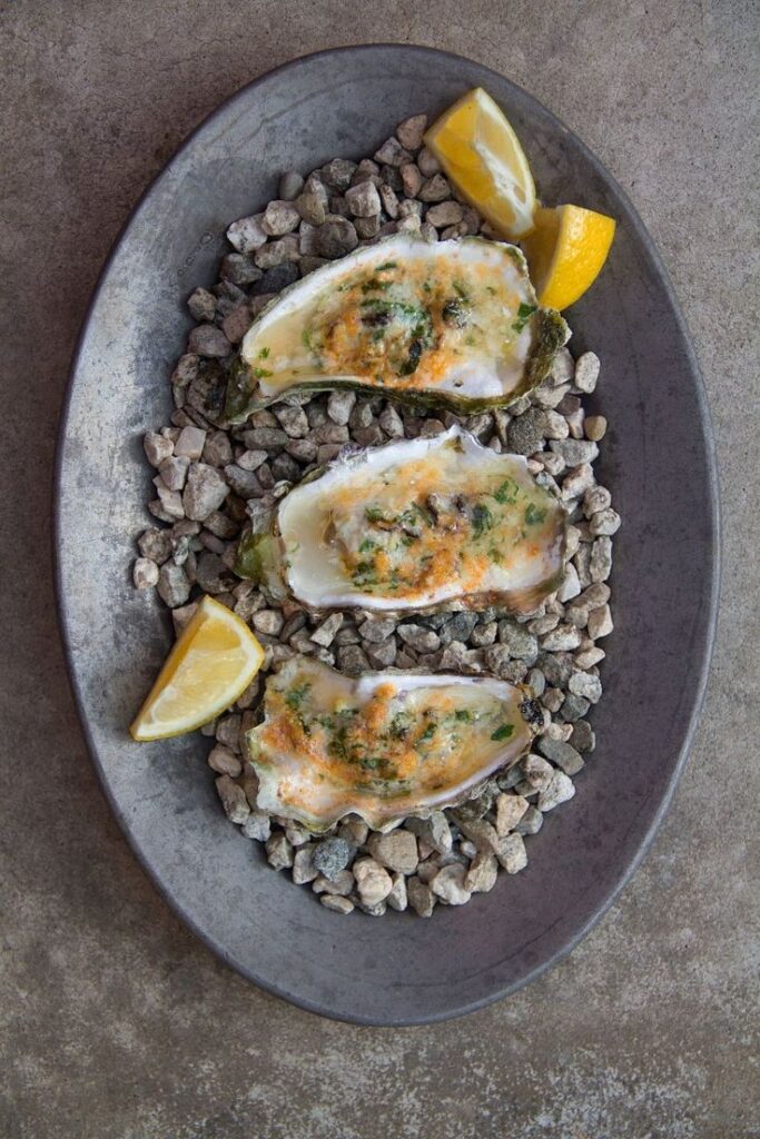 Grilled oysters with lemon wedges on a metal plate filled with pebbles.