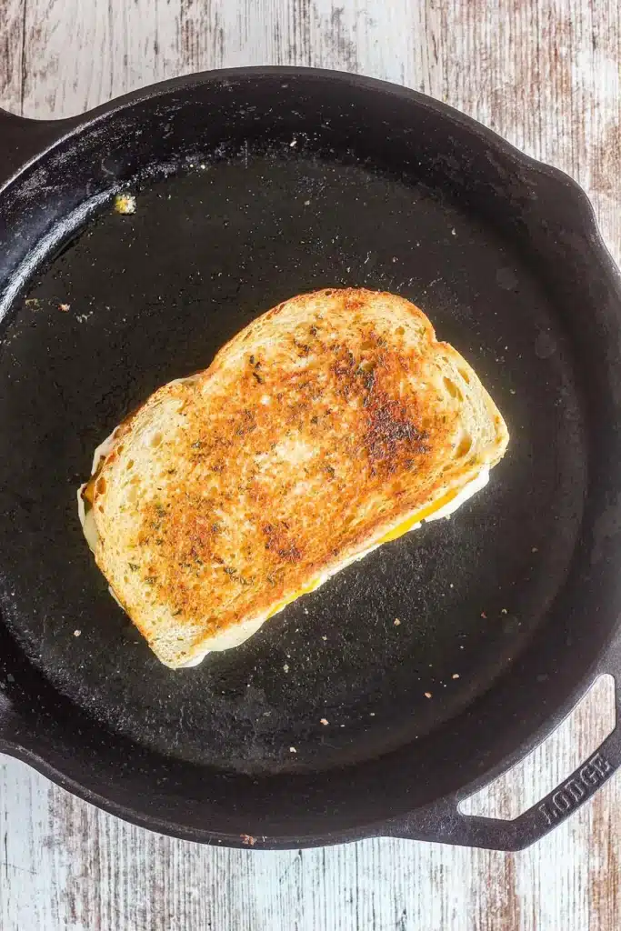 Grilled cheese sandwich toasting in a cast-iron skillet on a rustic wood surface.